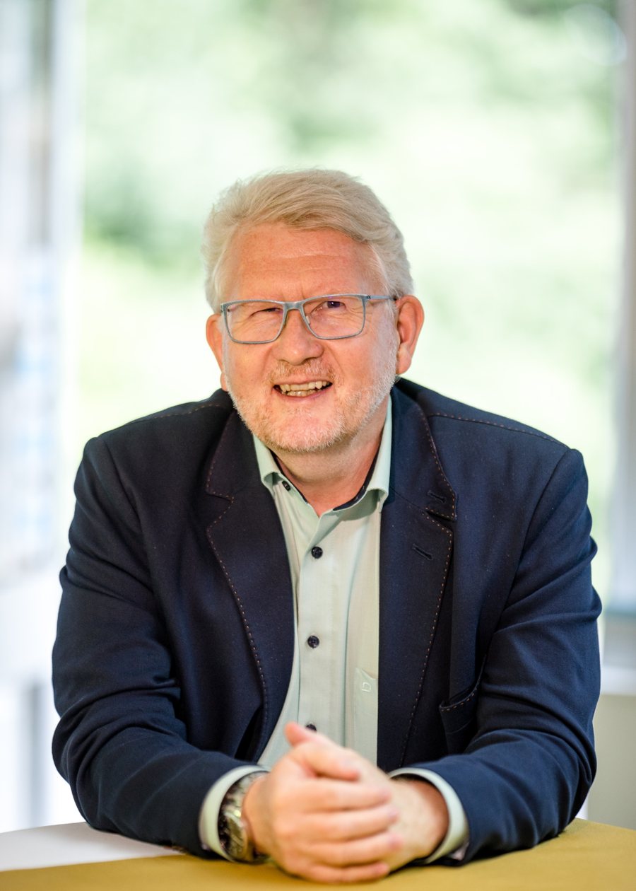 Detlef Gumze sitzt lächelnd am Tisch, die Hände gefaltet, helles Licht